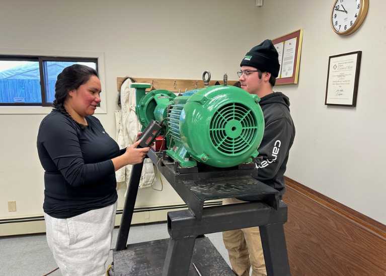 Two people work beside a green motor and pump assembly in a training room. One holds a measuring tool. This is an illustrative MRC library image, not a photograph of the Merritt cohort.