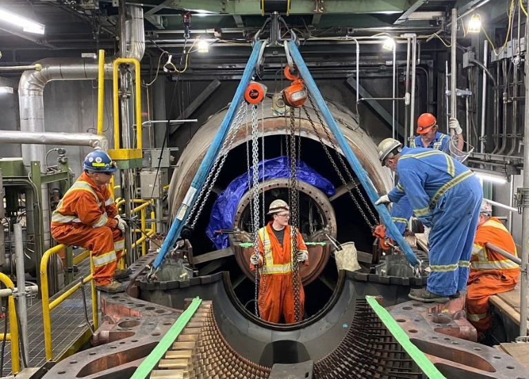 Five workers around an open turbine casing, with lifting chains and blue slings above the equipment.